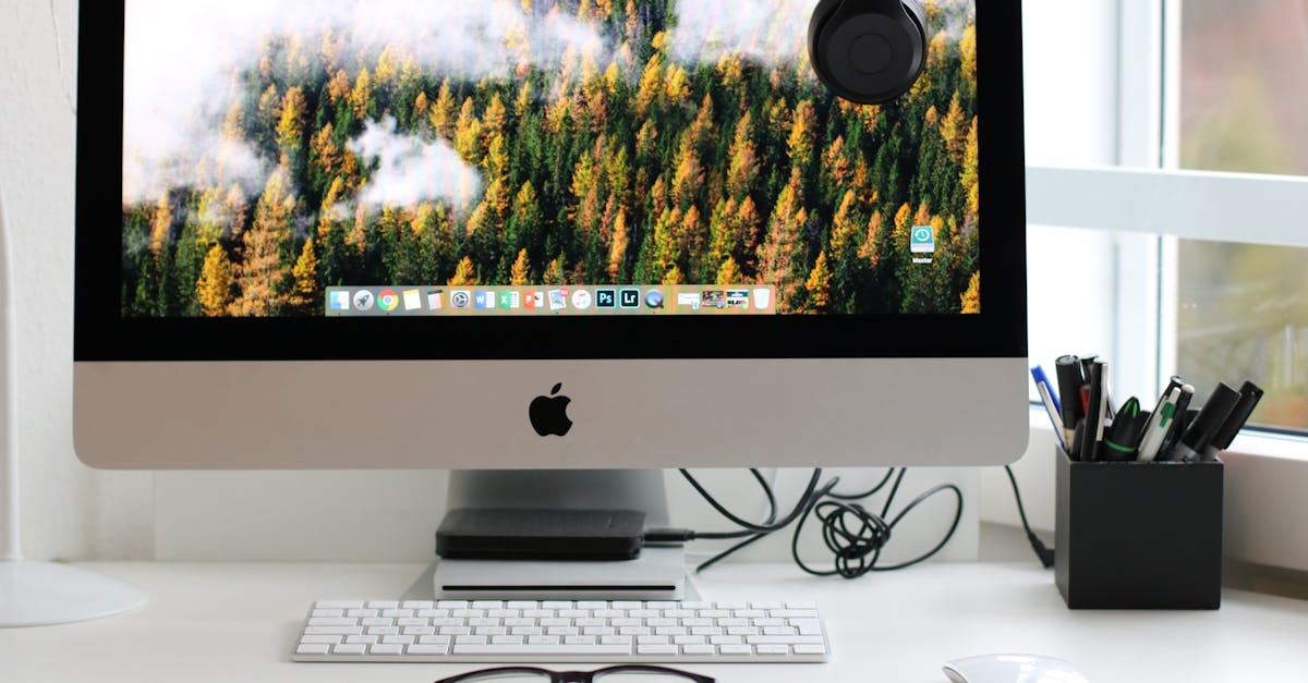 iMac desktop monitor displaying a vibrant forest landscape, with a wireless keyboard and glasses on a white desk, showcasing a modern home office setup ideal for productivity and design work.