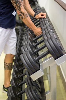 A man with tattoos selects dumbbells from a rack in a gym setting, focused on strength training.