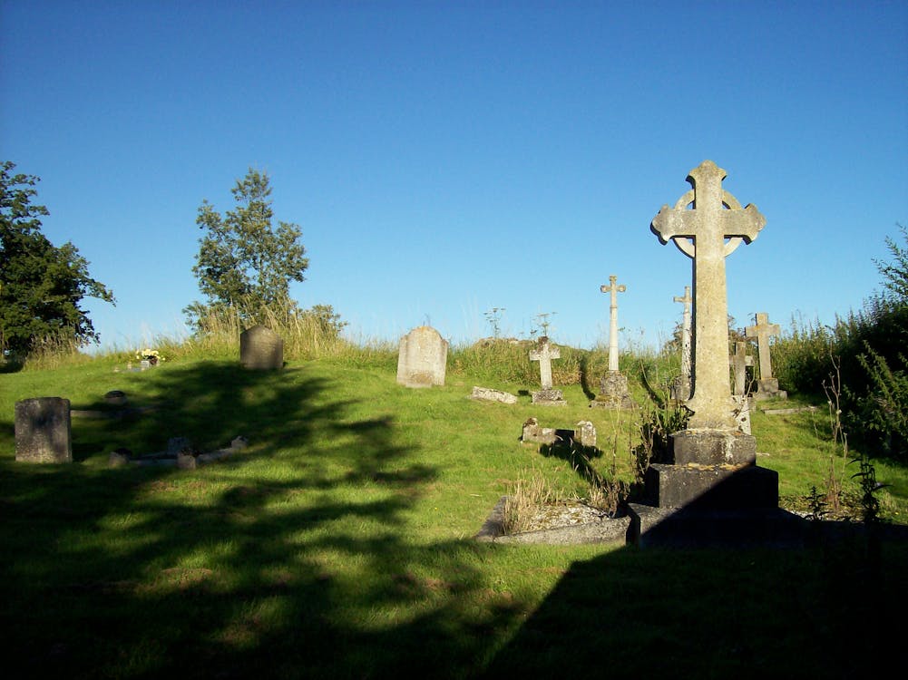  An overgrown graveyard with a large Celtic cross in the foreground and other gravestones in the background.