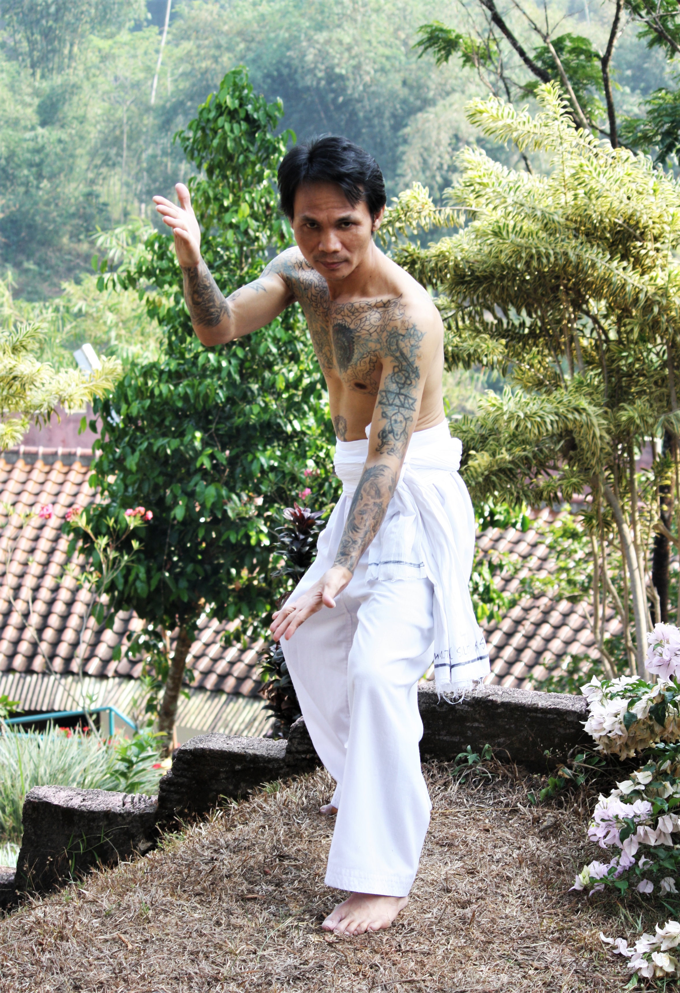 Topless man with tattoos in a martial arts pose in a lush, outdoor setting.