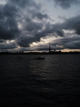 A moody silhouette of skyline and boats at sunset, with dramatic clouds over a dark ocean.