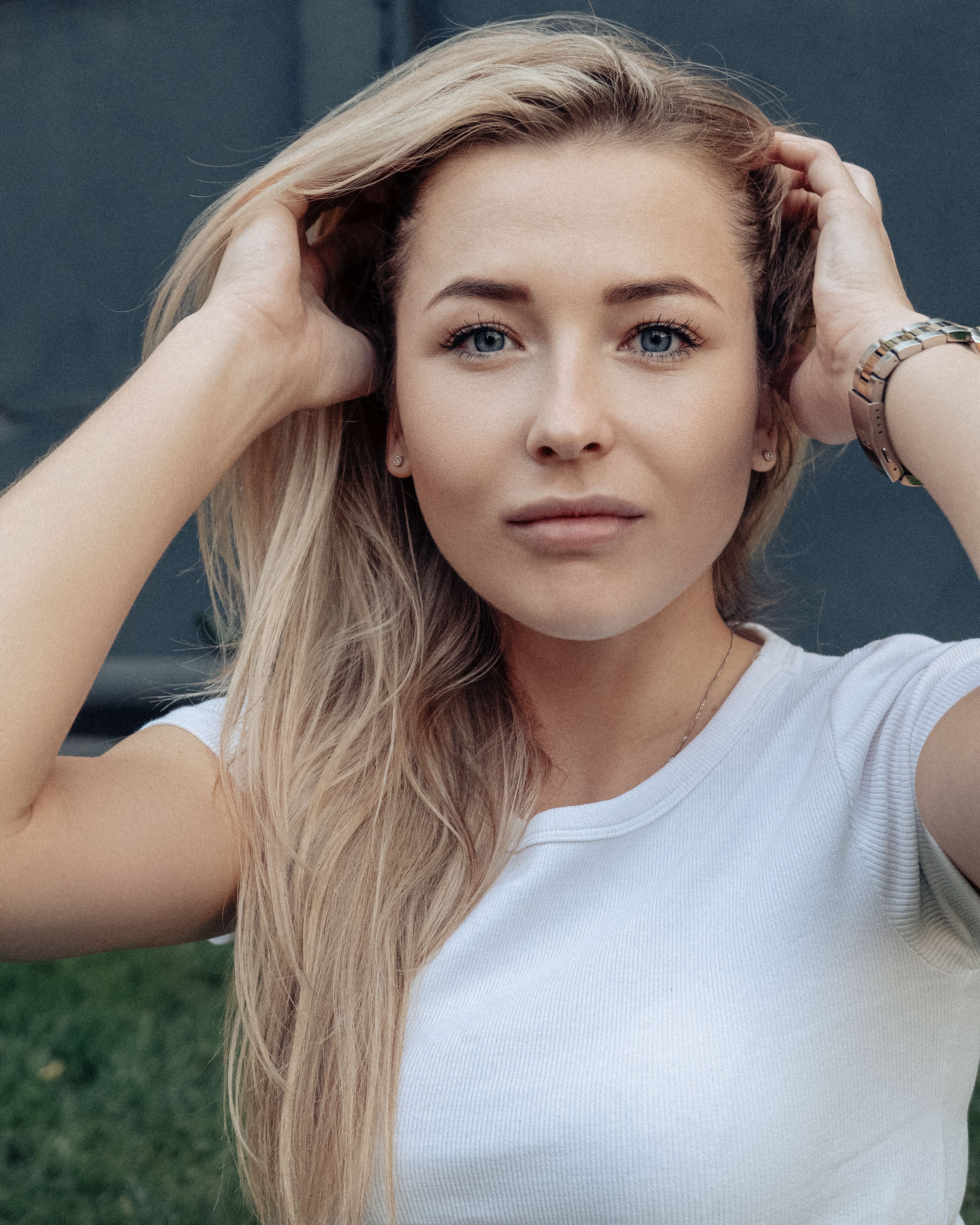 Portrait of a blonde woman outdoors, looking directly at the camera, hands touching her hair.