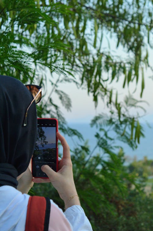 Person in Black Hijab Holding Black and Red Smartphone Taking Photo of Green Trees