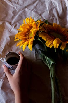 A close-up of a hand holding a coffee cup beside vibrant sunflowers on crumpled paper.