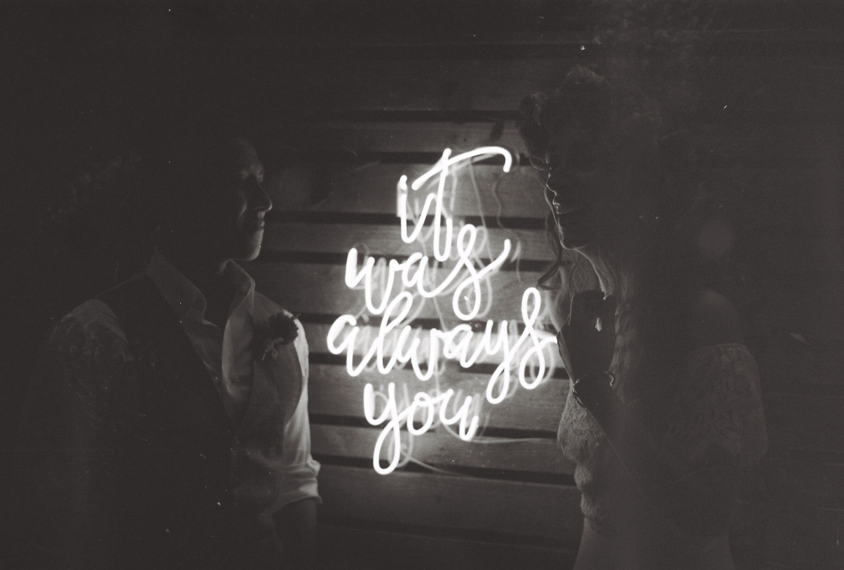 Black and white photo capturing a romantic moment in front of a neon sign reading 'it was always you'.