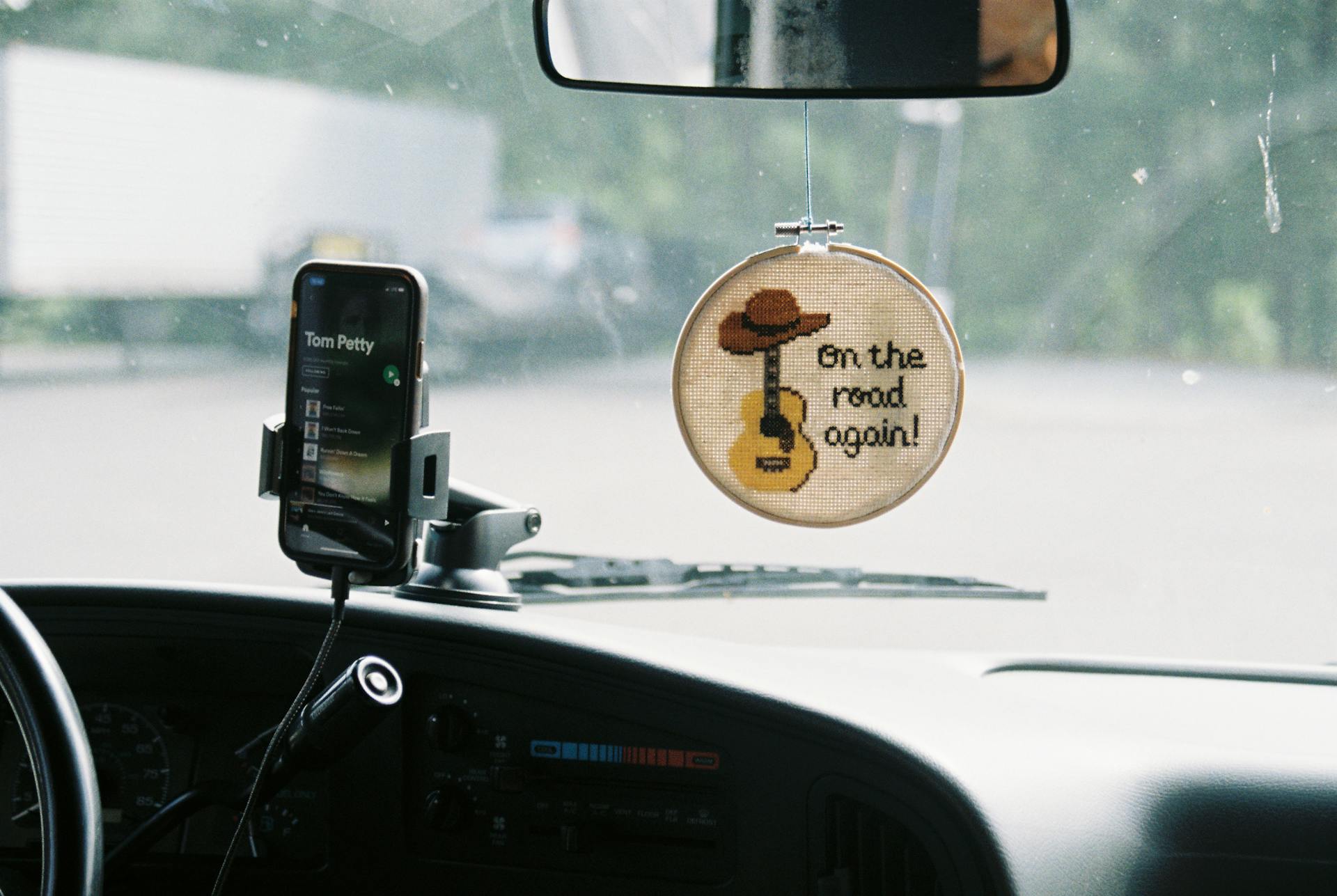 Dashboard view with phone playing music and cross-stitch hanging decor.