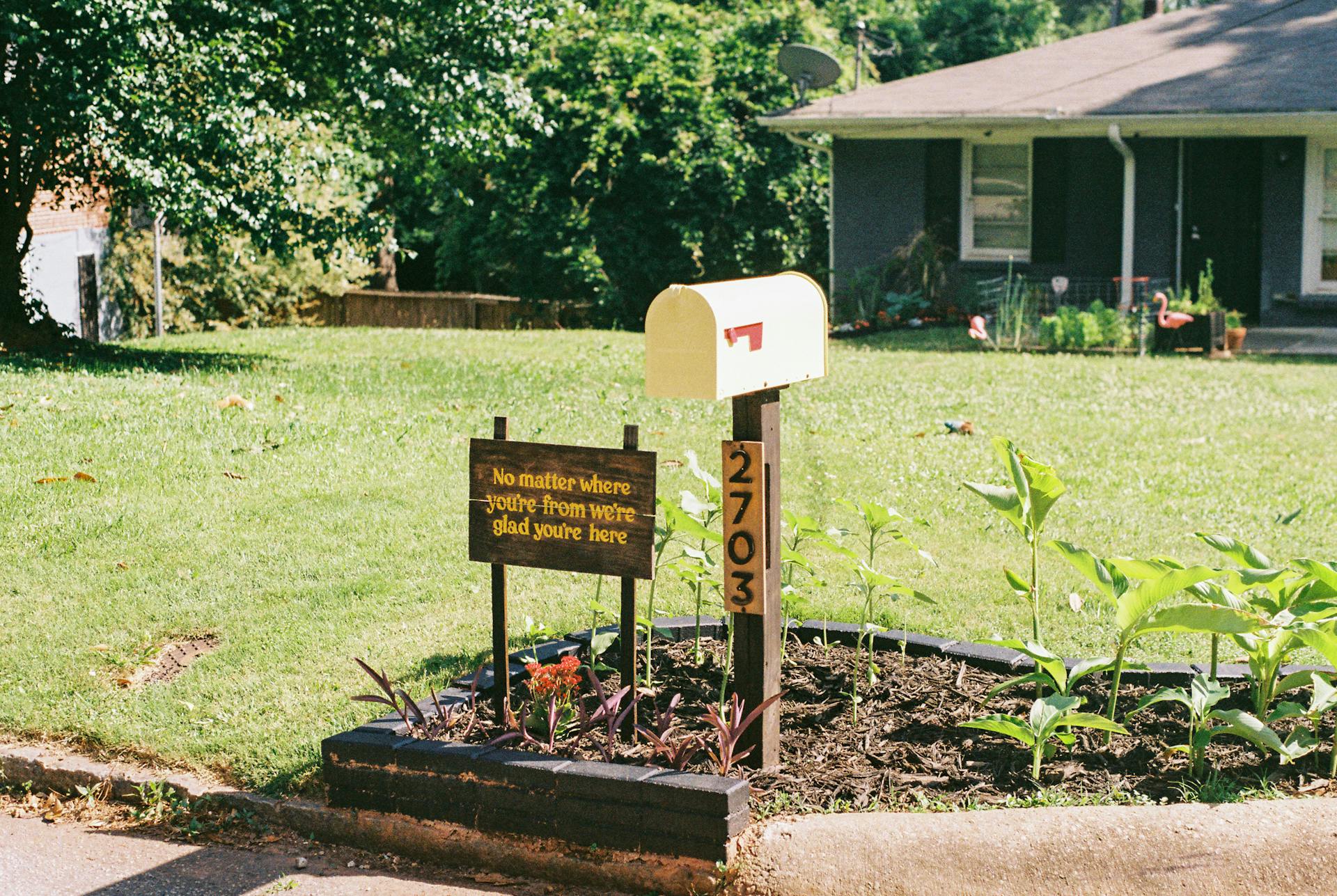 Yellow mailbox with a warm welcoming sign and plants in a suburban front yard.