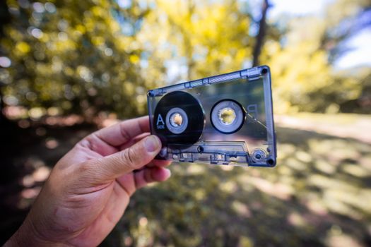 A close-up of a hand holding a vintage cassette tape outdoors with a blurred background.