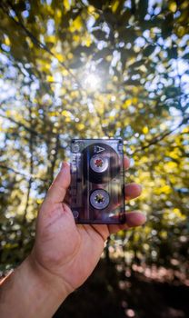 A hand holding a cassette tape against a background of sunlight streaming through trees.