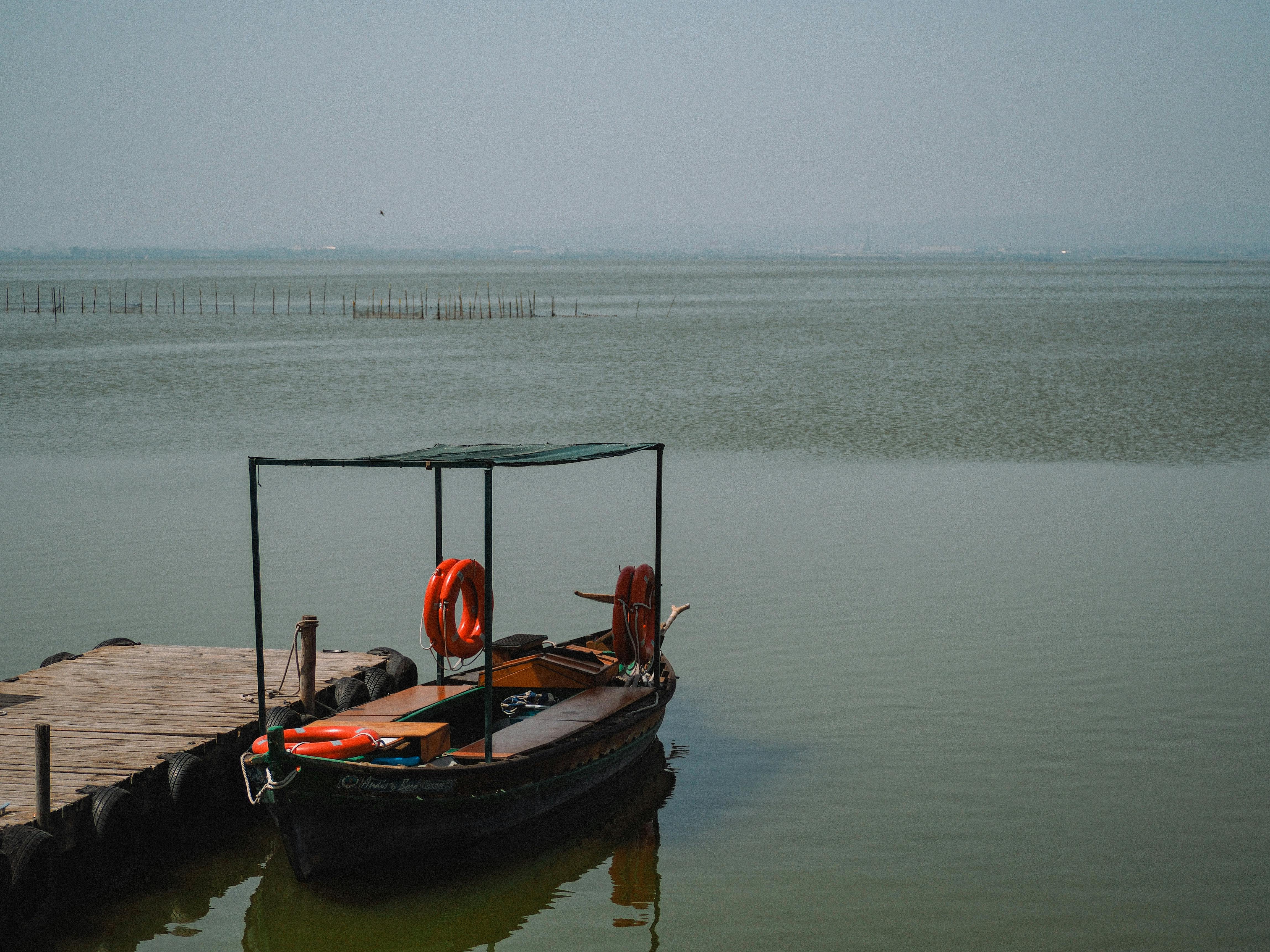 Free Peaceful scene of a small boat docked by a wooden pier on a calm lake under cloudy skies. Stock Photo