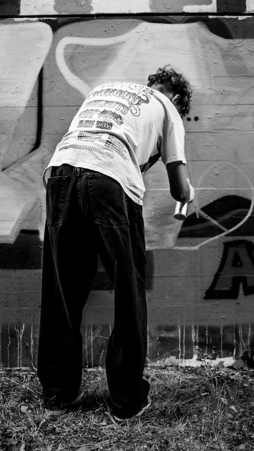 Grayscale Photo of a Street Artist Spray Painting a Wall