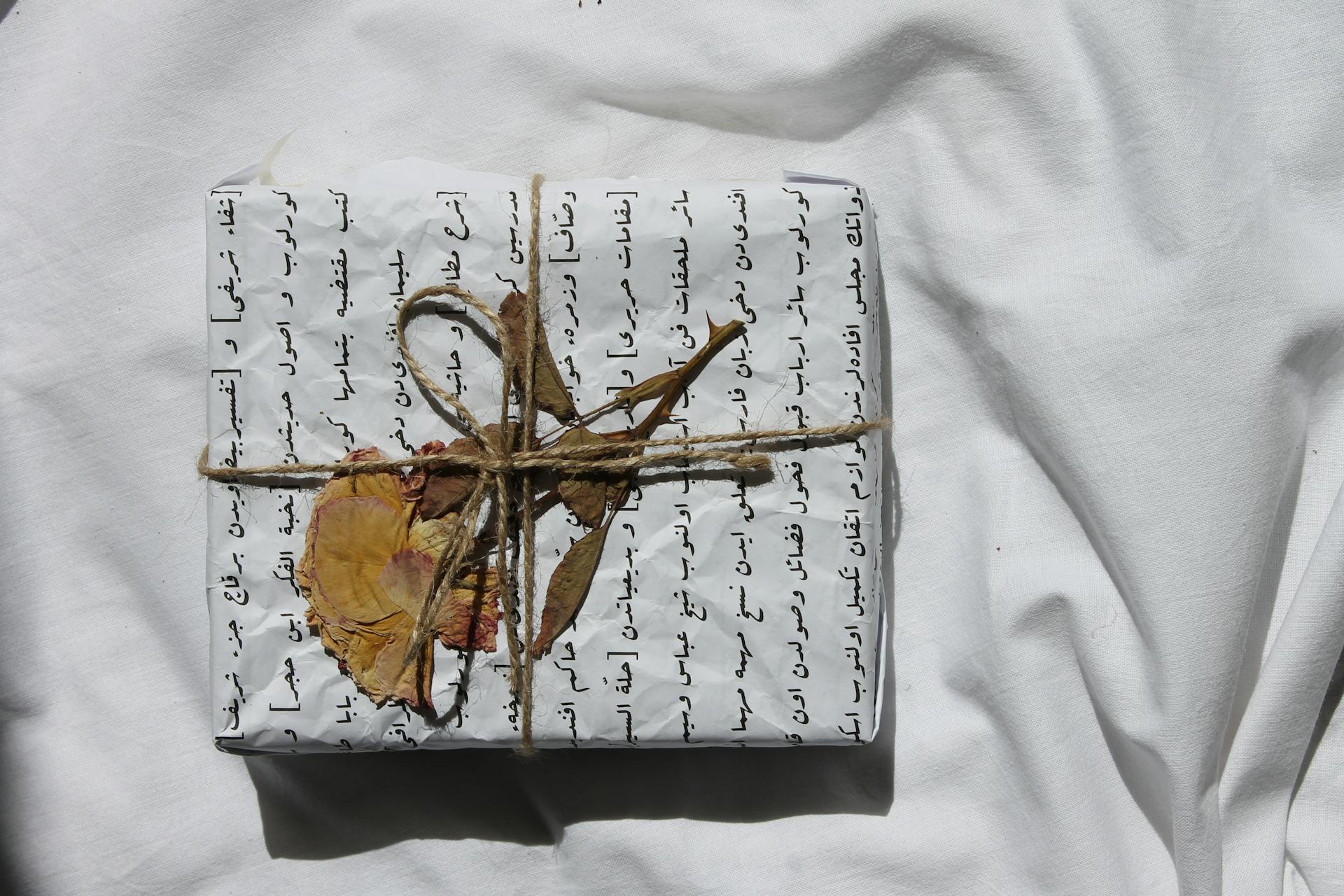 A gift wrapped in Arabic script paper, adorned with a dried flower and twine on white fabric.