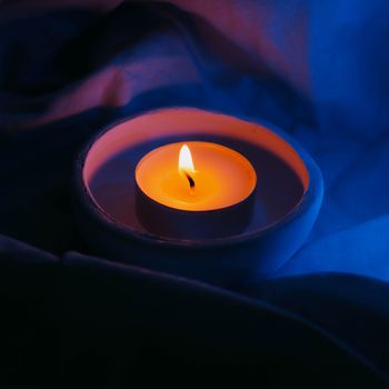 Close-up of a burning tealight candle in a dark, moody setting with contrasting blue and orange tones.