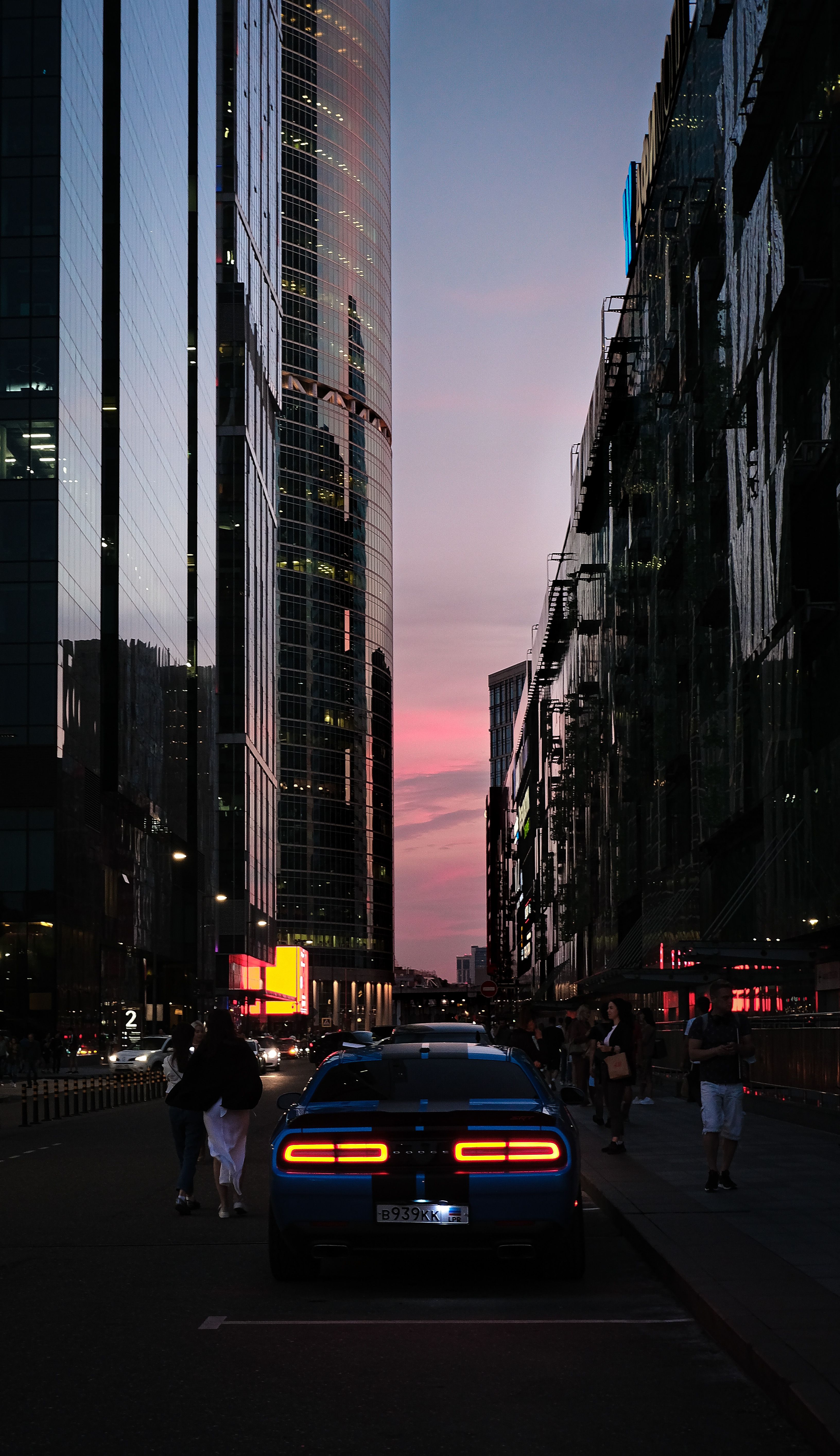 Blue car on city street at dusk with people and towering skyscrapers in twilight.