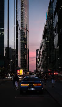 Blue car on city street at dusk with people and towering skyscrapers in twilight.