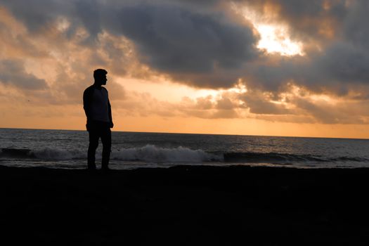 Silhouette of a man standing on a beach as the sun rises, casting a warm glow over the ocean.