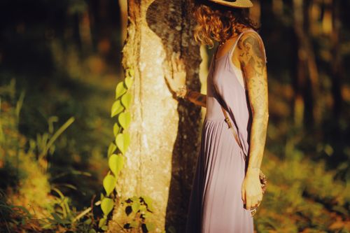 Free A tattooed woman in a purple dress standing by a tree trunk in a forest during sunset. Stock Photo