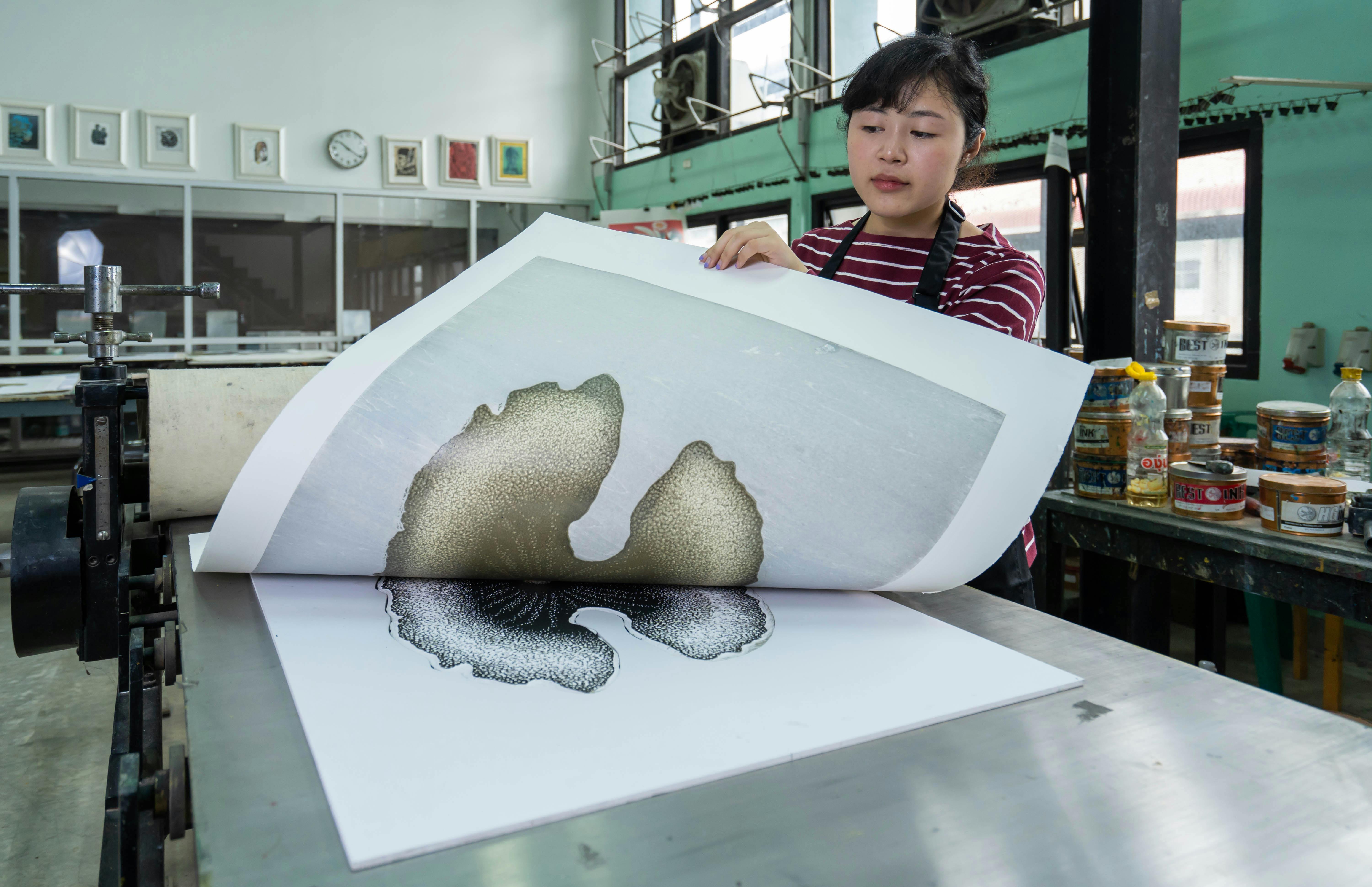 A woman inspects a freshly printed design in a modern printmaking studio, surrounded by printing tools and equipment.