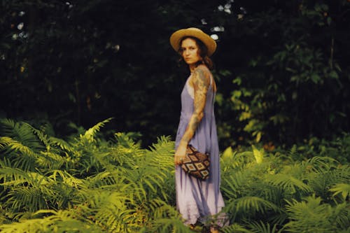 A Woman in Ling Dress Wearing Straw Hat while Standing on the Ground Surrounded by Green Plants while Looking at the Camera