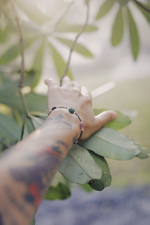 Free Close-up of a tattooed arm with a bracelet touching green leaves outdoors. Stock Photo