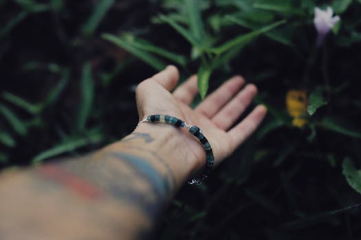 A tattooed hand with a bracelet extended outdoors against a lush leafy background.