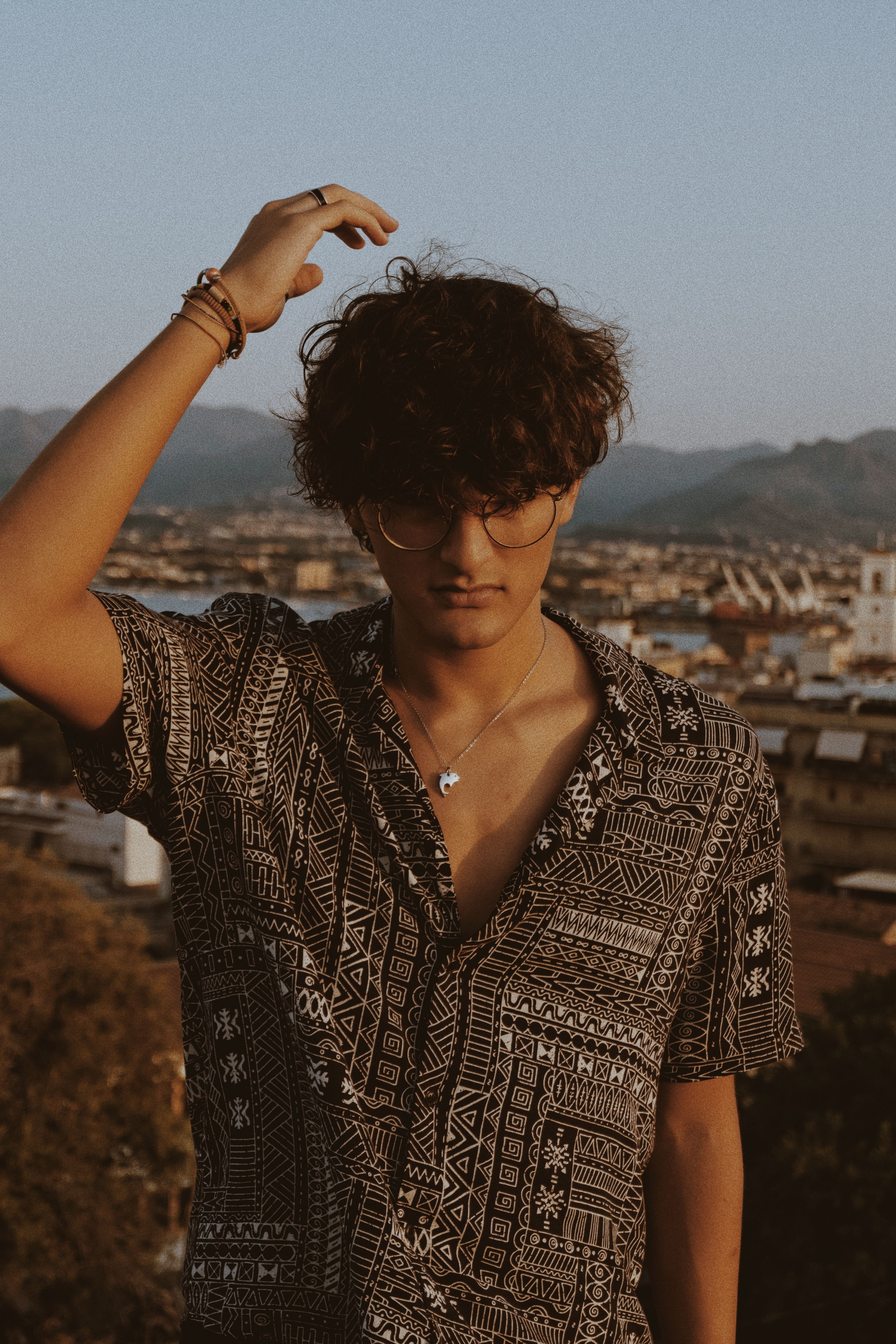 Stylish young man with glasses and curly hair poses against a vibrant city skyline during a warm evening.