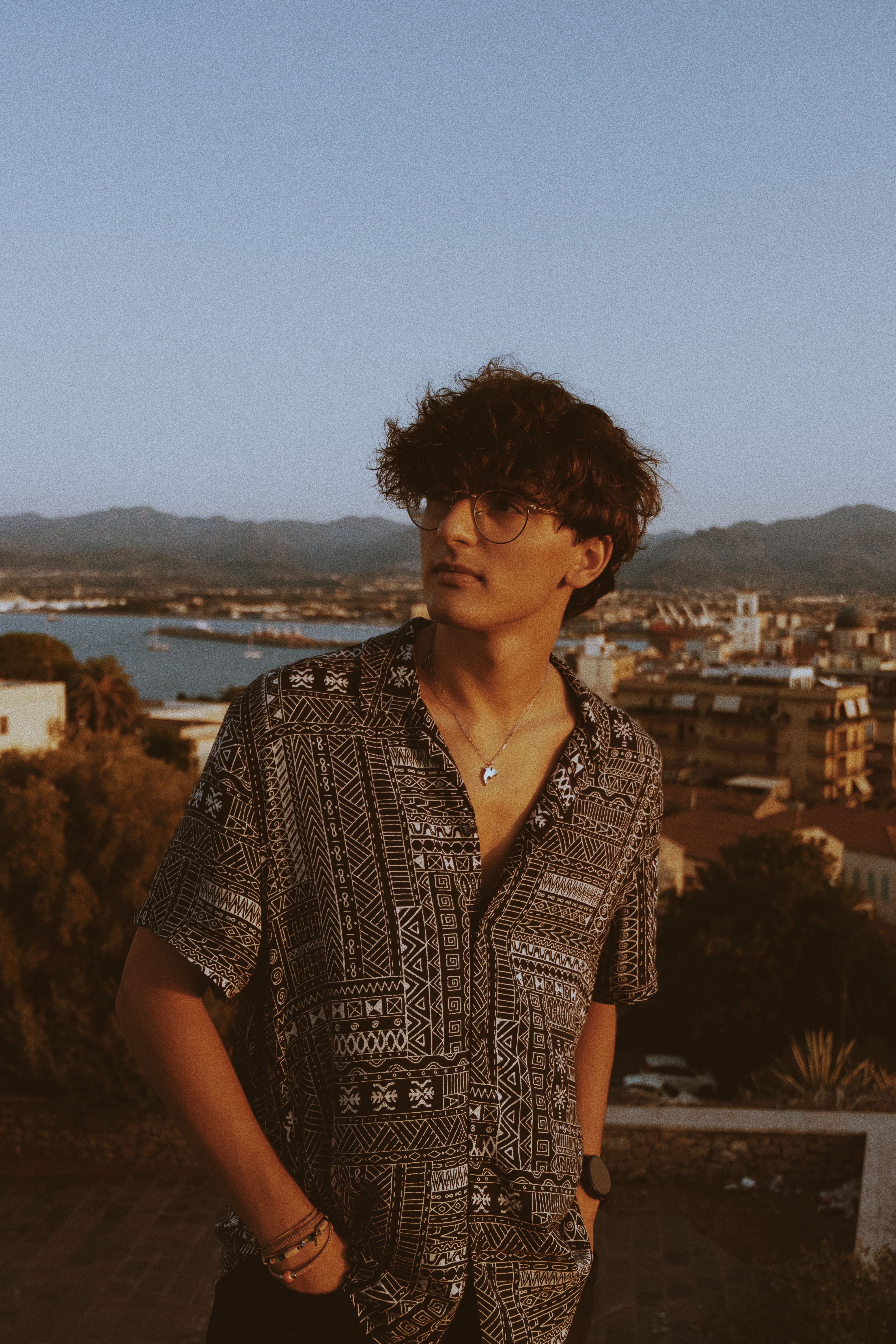 Young man with curly hair and glasses gazes over a city skyline at sunset, wearing a patterned shirt.
