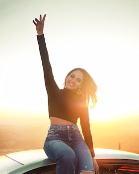 Smiling young woman in stylish outfit joyfully raises arm sitting outdoors at sunset.