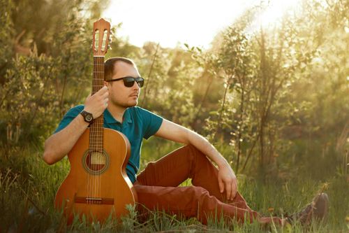Free A man sitting in grass holding a guitar, enjoying a sunny day outdoors. Stock Photo