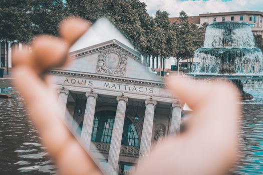 A handheld mirror reflects the historic Kurhaus Wiesbaden facade near a vibrant fountain.