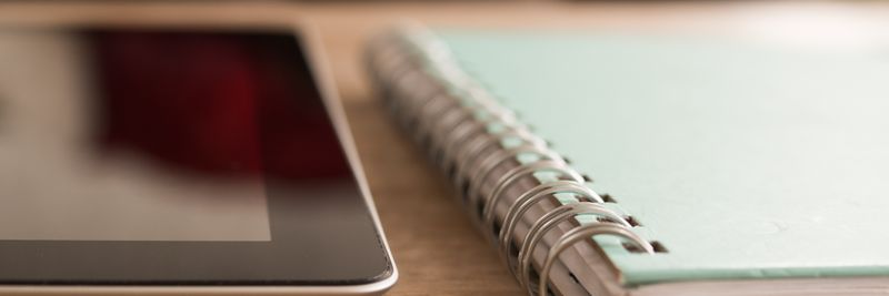 A close-up photo of a tablet alongside a spiral notebook on a wooden desk.