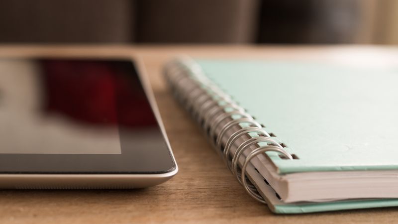 A close-up photo of a tablet alongside a spiral notebook on a wooden desk.