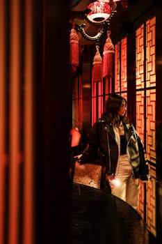 Fashionable woman in black leather jacket posing in a glamorous red-lit interior with intricate decor.