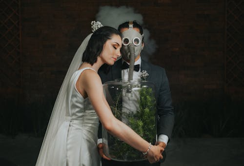 Bride Holding a Huge Glass Jar wit Green Plants for his Groom Wearing a Gas Mask
