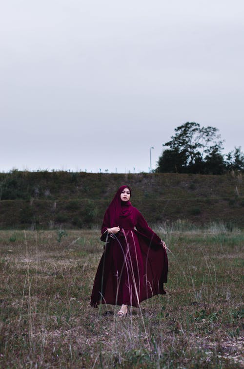 Free A Woman Wearing Red Hijab Standing on a Grass Field while Seriously Looking at the Camera Stock Photo