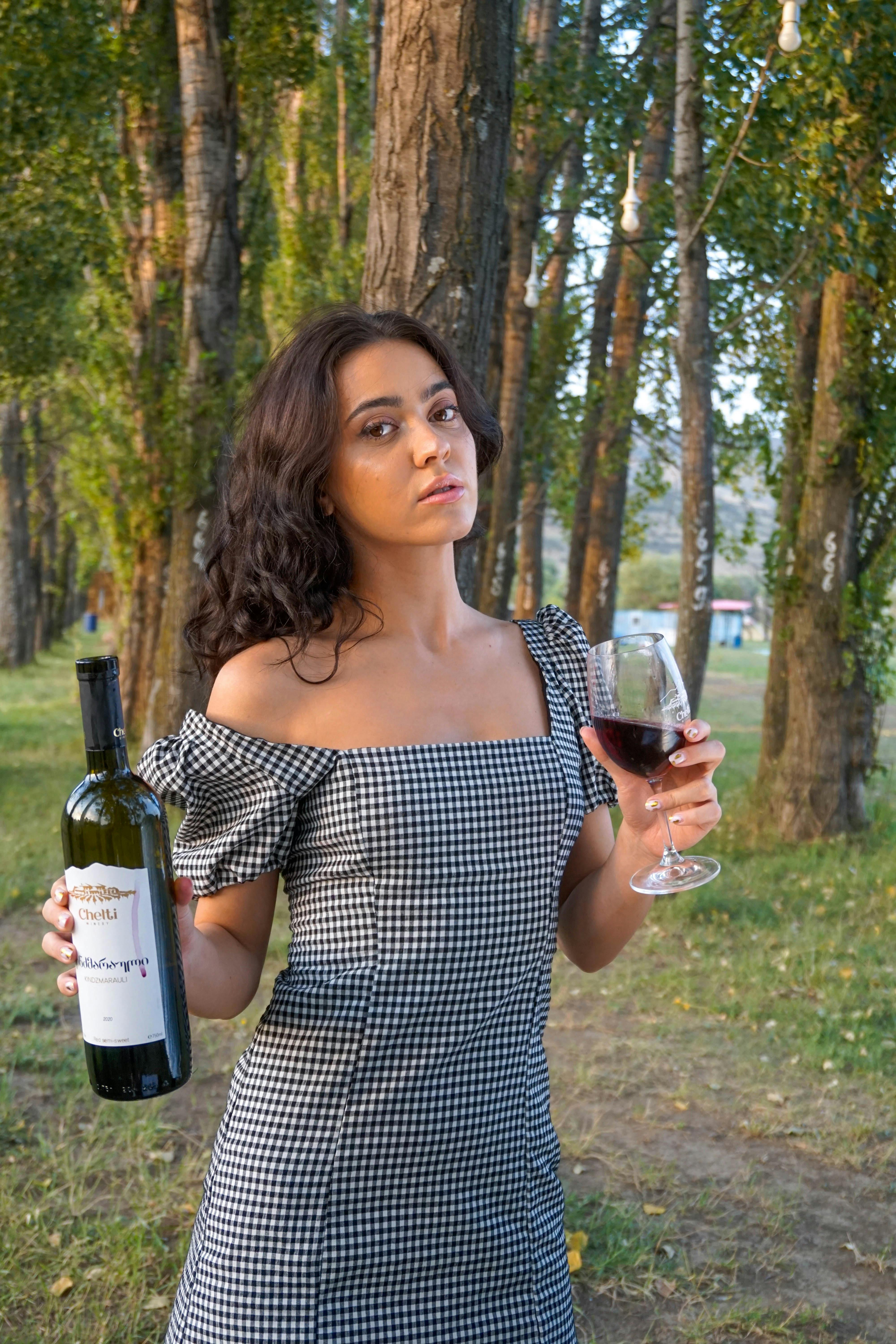 A woman in a checkered dress holds a bottle and glass of red wine outdoors among the trees.