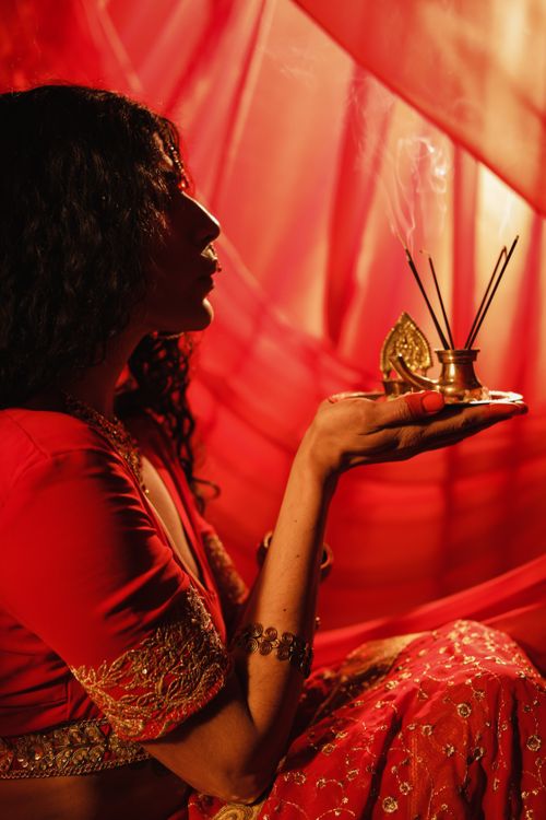 Free Indian woman in traditional attire holding incense, captured in a serene side profile with vibrant red background. Stock Photo