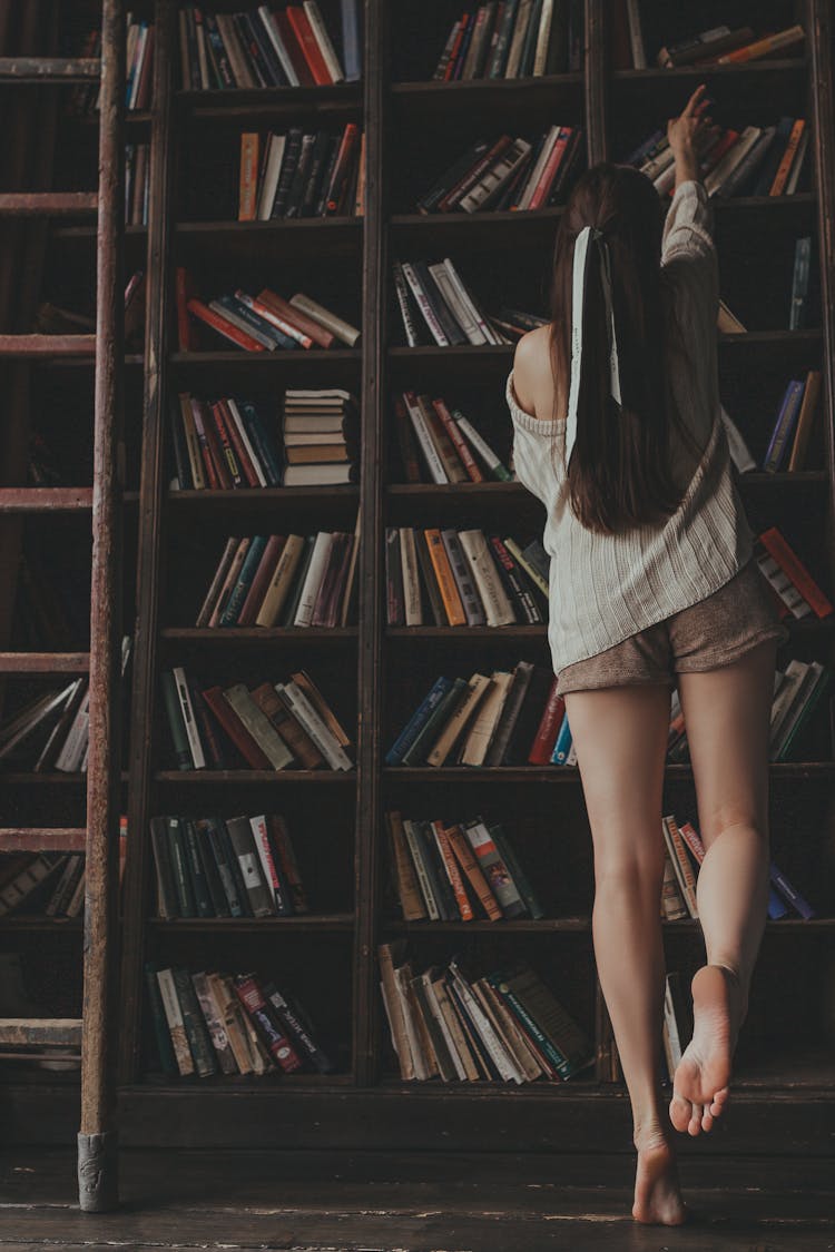 A Back View Of A Woman Getting A Book From The Shelf