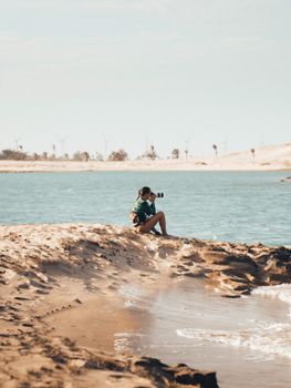 A woman sits on a sandy beach, taking photos of the serene coastal landscape with wind turbines in the background.