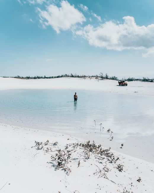 A lone person stands in a serene lagoon surrounded by sandy dunes under a clear blue sky in Cabo Frio, Brazil.
