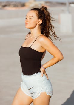 A woman stands outdoors in a black top and shorts, enjoying the sun with closed eyes.