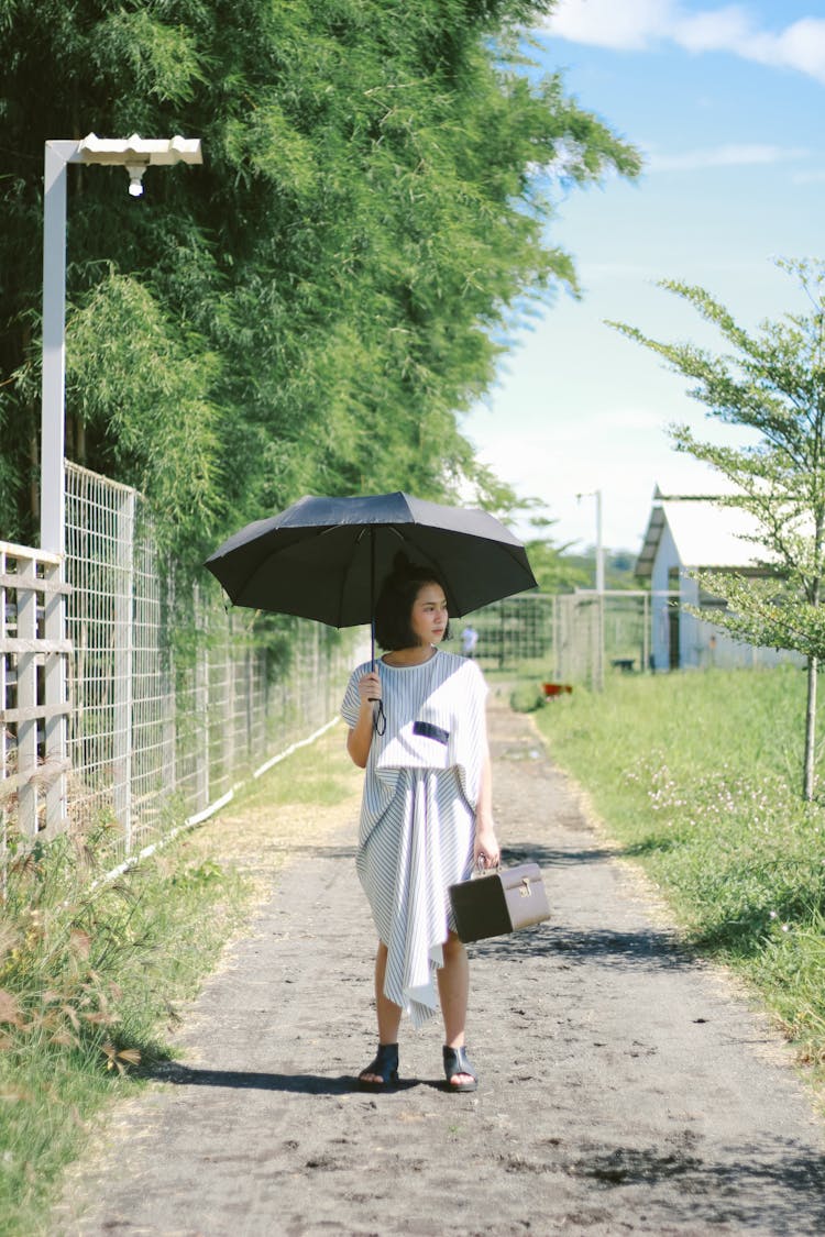 Woman In A Frilly Dress Walking Under Black Umbrella On A Footpath Along A Fence