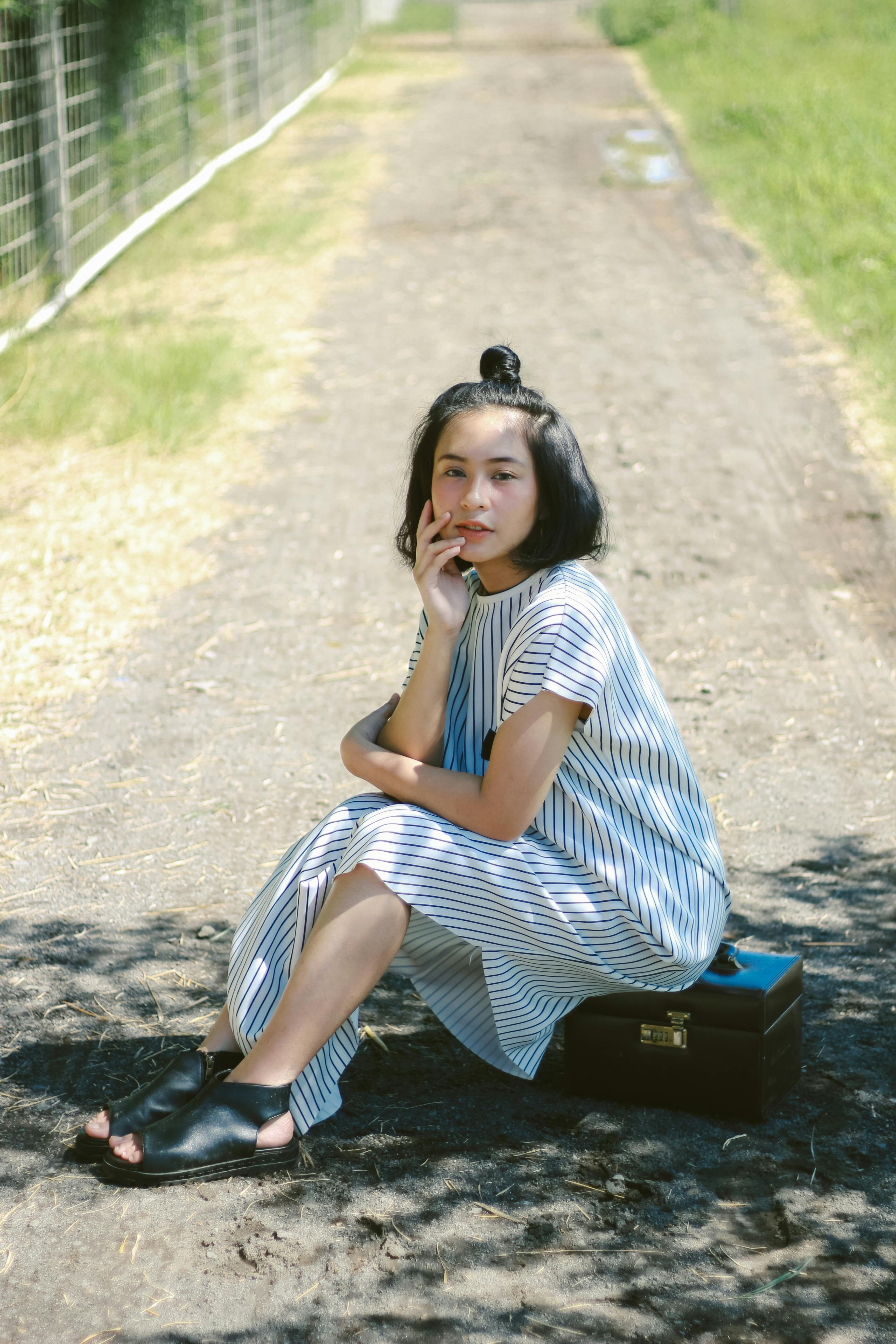 Free Asian woman in striped dress sitting on a path, posing thoughtfully outdoors on a sunny day. Stock Photo