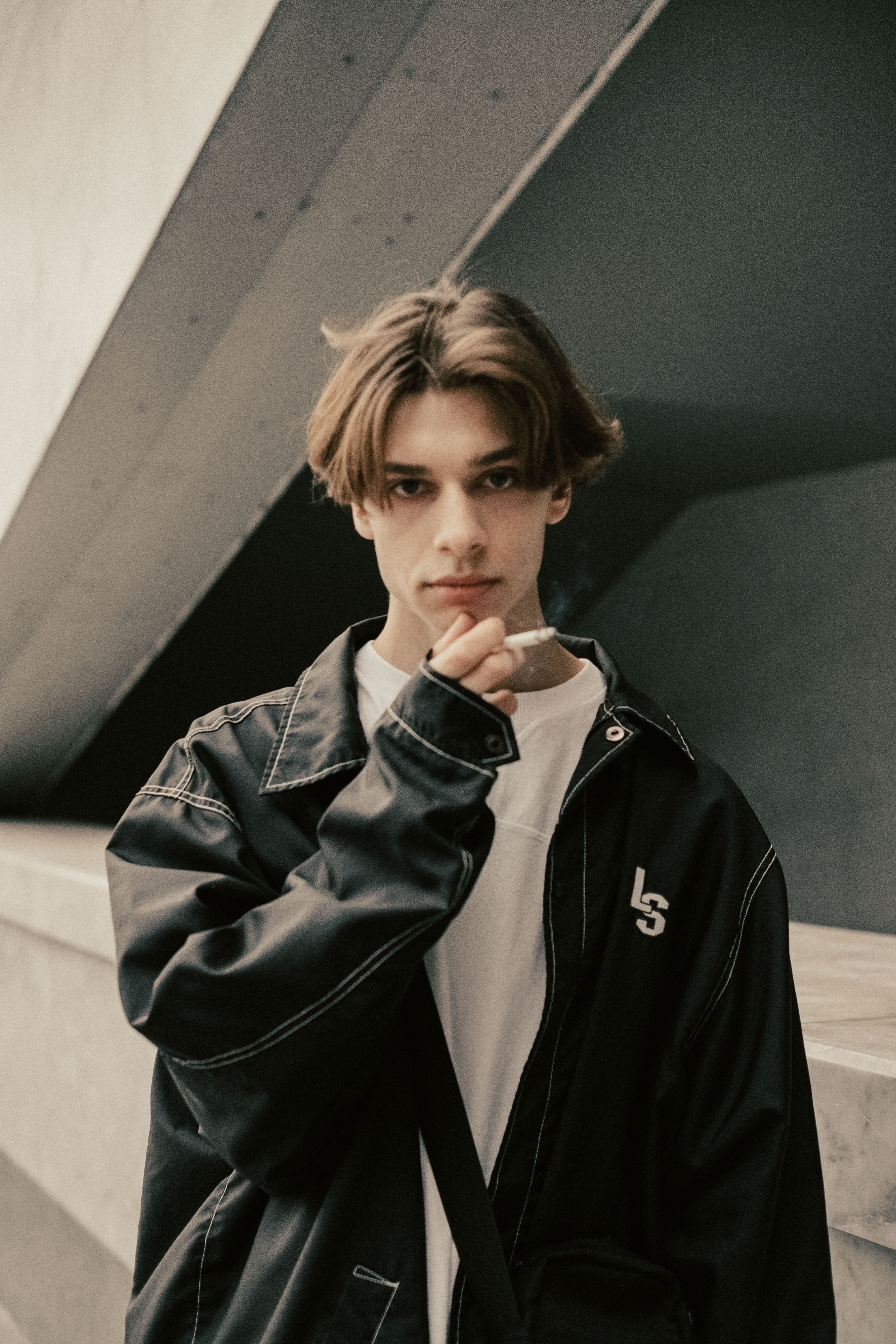 Young man in a casual jacket holding a cigarette with architectural backdrop.