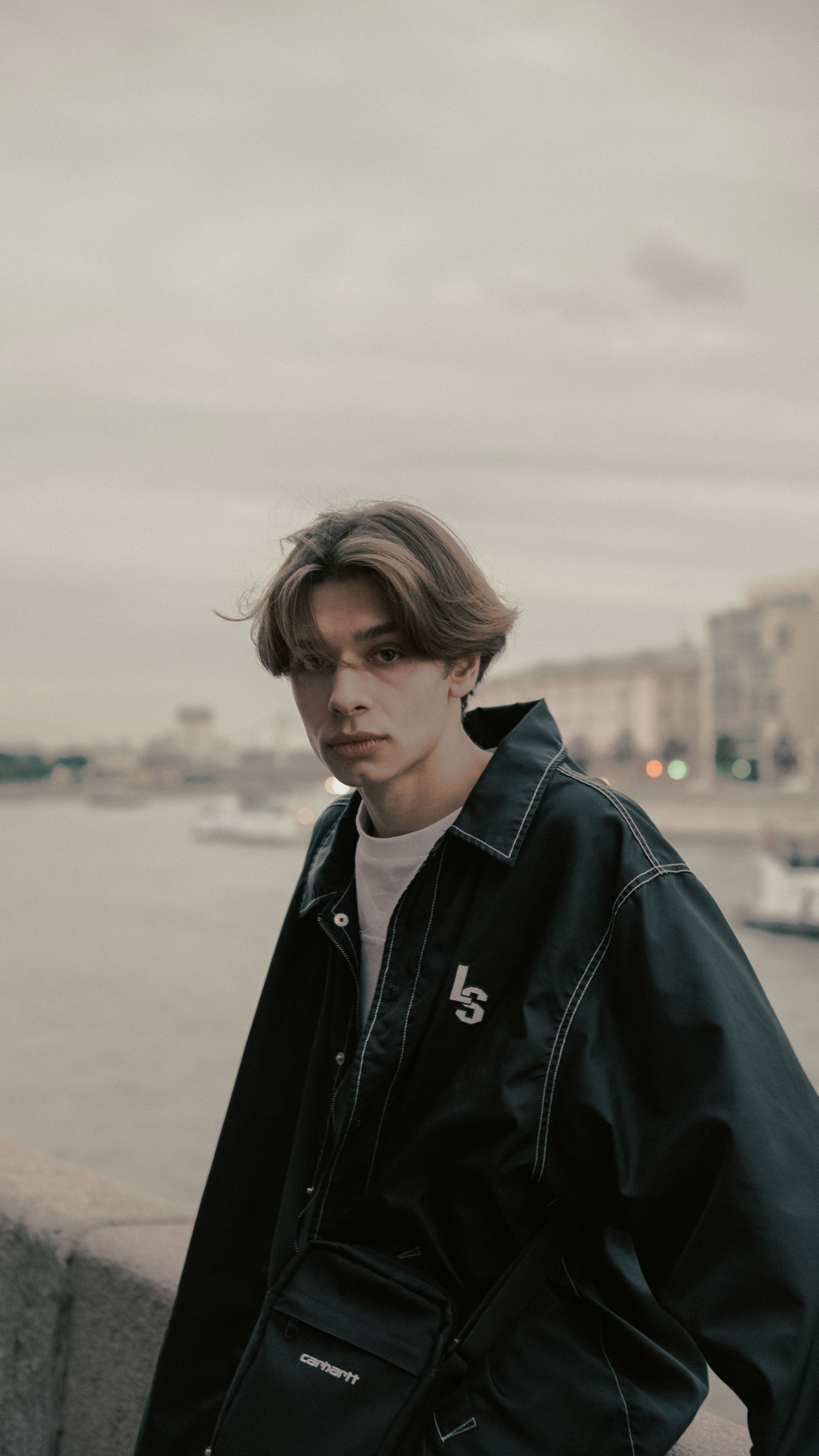 A young man in a black jacket posing by a waterfront with cityscape background.