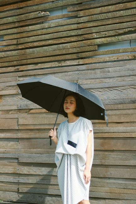 Stylish woman holding an umbrella, posing gracefully against a rustic wooden wall outdoors.