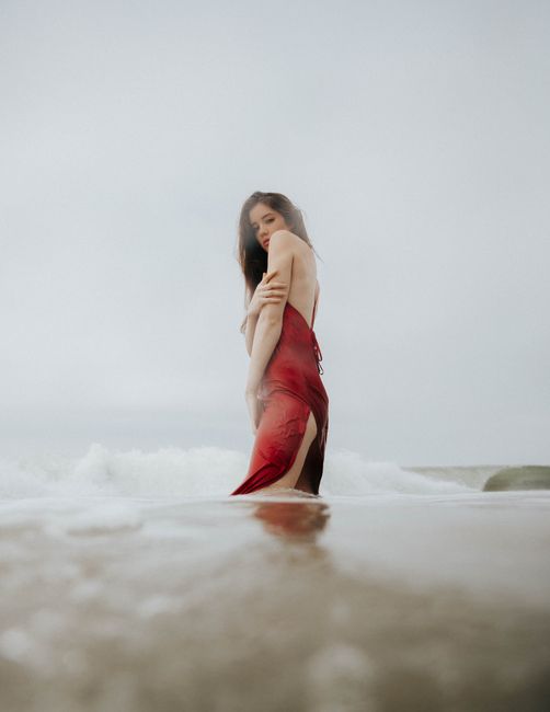 A woman in a red dress kneels at the ocean shore, gazing at the camera with soft waves around her.
