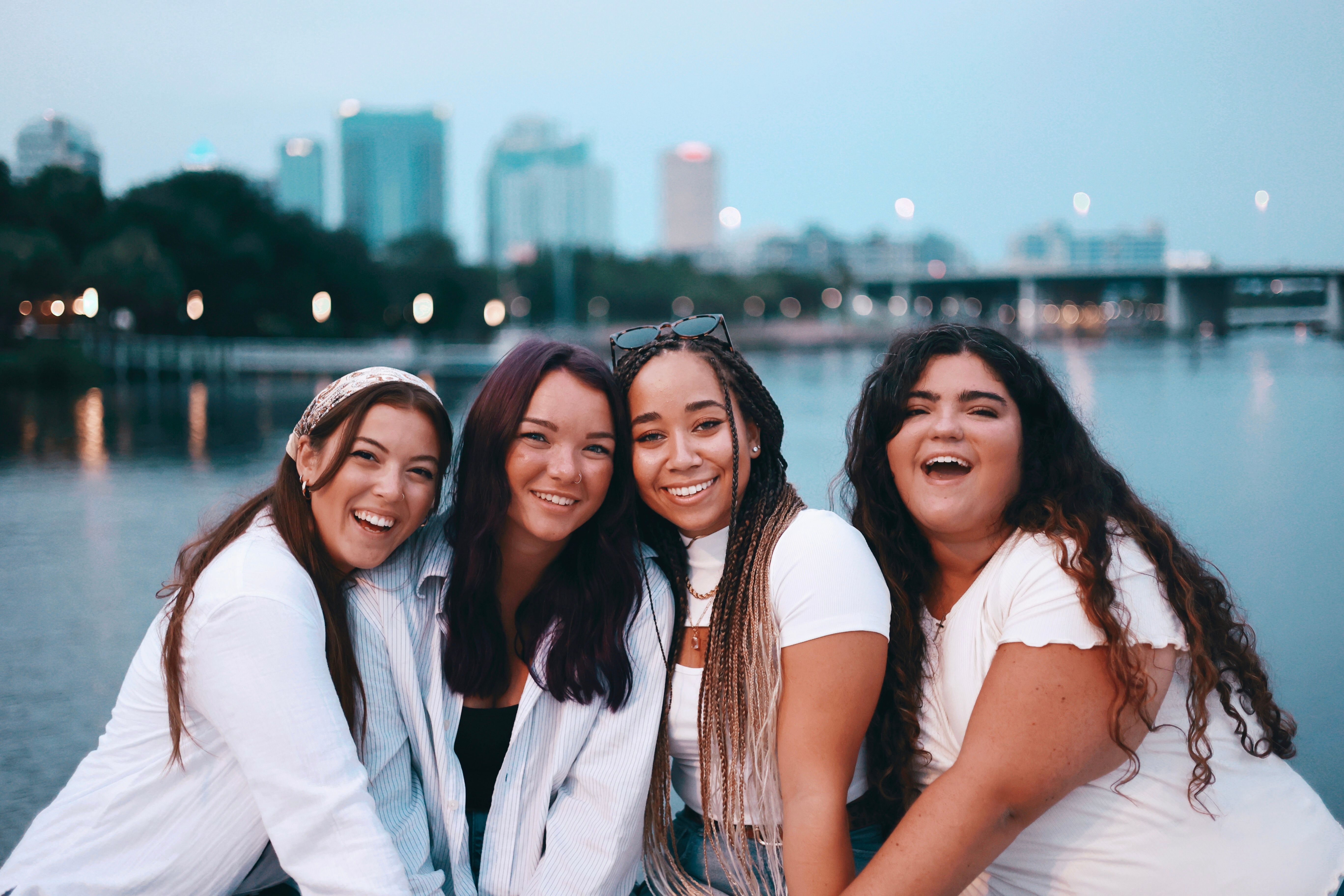Four women smiling together by the city waterfront during the day.