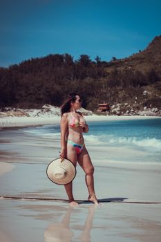 Woman in a vibrant bikini and sun hat enjoying a sunny day on a tranquil beach.