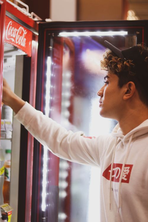 Free A young man in a white hoodie reaching into a Coca-Cola-branded refrigerator inside a store. Stock Photo